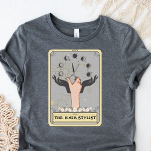 Puede incluir: Una camiseta gris con un gr&aacute;fico de un dise&ntilde;o de carta de tarot que presenta una mano sosteniendo tijeras y el texto "THE HAIR STYLIST".