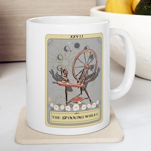 May include: White ceramic mug with a design of a tarot card depicting a spinning wheel with a gray background and the text "XXVII THE SPINNING WHEEL".