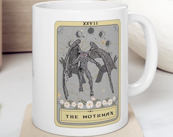 May include: White ceramic mug with a graphic of a creature with wings and red eyes. The graphic is in a tarot card style with the text "XXVII THE MOTHMAN".