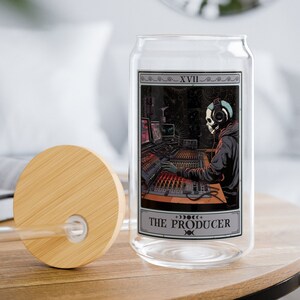 May include: A clear glass can with a bamboo lid and a straw. The can features a tarot card design with a skull wearing headphones and sitting at a soundboard. The card reads "XVII THE PRODUCER".