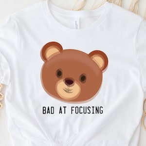 May include: White t-shirt with a brown cartoon bear face and the text "Bad at Focusing" printed on the front.