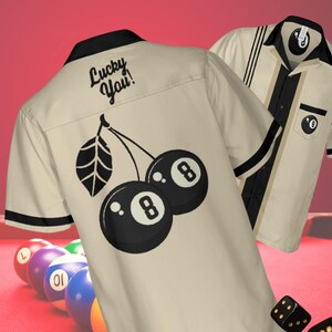 以下が含まれることがあります： 黒のストライプと黒の8ボールチェリーのデザインが施されたベージュのボウリングシャツ。シャツの背中には「Lucky You!」の文字が書かれている。