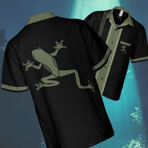 May include: Two black button-down shirts with a frog graphic. One shirt features a large olive green frog on the back. The other shirt has olive green stripes and a small frog on the pocket. Both shirts have olive green accents.