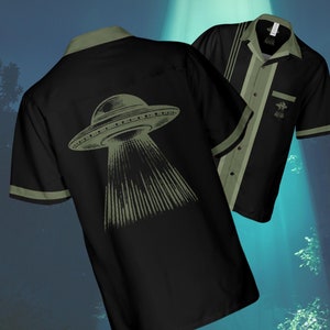 Könnte beinhalten: Ein schwarzes und grünes Bowlinghemd mit einer Vintage-Illustration einer fliegenden Untertasse auf der Rückseite. Das Hemd hat einen Knopfkrag und kurze Ärmel.