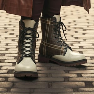 May include: Lace-up combat boots with a cream-colored toe and a brown plaid pattern on the upper. The boots have black laces and a brown sole. The boots are worn on a brick surface.