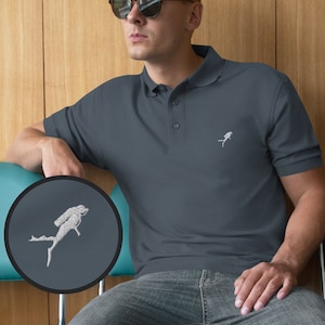 May include: A dark blue polo shirt with a white embroidered scuba diver design on the left chest. The shirt has a button-up collar and short sleeves.