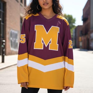 May include: A maroon and gold hockey jersey featuring a large white-outlined gold "M" on the front. The jersey has gold sleeves with white and gold chevron stripes, and the numbers "5" and "2" are on the sleeves.