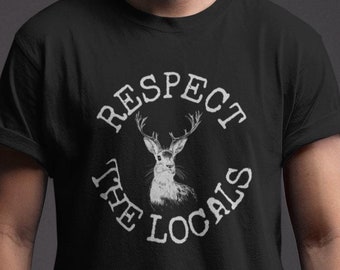 Può includere: Maglietta nera con una grafica circolare bianca che dice "Respect the Locals" con una testa di cervo all'interno del cerchio.