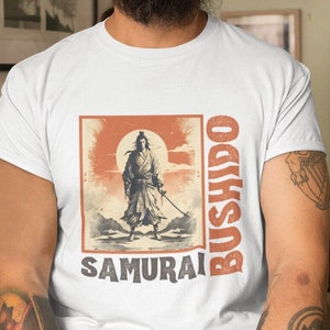 May include: White t-shirt featuring a vintage-style illustration of a samurai warrior with a sword, set against a rising sun. The words "BUSHIDO" and "SAMURAI" are printed in orange. The shirt is a classic crew neck.