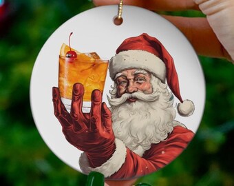 Può includere: Un ornamento natalizio in ceramica bianca con un Babbo Natale dei cartoni animati che tiene un bicchiere di cocktail arancione con una ciliegia come guarnizione. L'ornamento è rotondo e ha un filo d'oro per appenderlo.