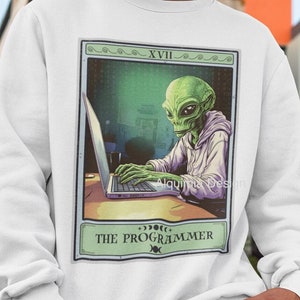 Puede incluir: Sudadera blanca con un extraterrestre verde que lleva una sudadera con capucha y escribe en un portátil. La imagen está en estilo de carta de tarot con el texto "XVIII THE PROGRAMMER" en la parte inferior.