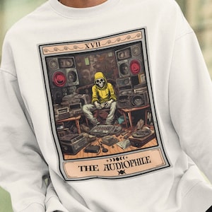 May include: White sweatshirt featuring a graphic print of a tarot card. The card is titled "The Audiophile" and depicts a skeleton wearing a yellow hoodie sitting in a room surrounded by audio equipment. The card number is XVII.