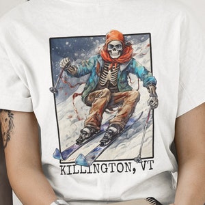 May include: White t-shirt with a graphic of a skeleton skiing down a snowy mountain. The graphic is in a rectangular frame with the text "KILLINGTON, VT" below it.