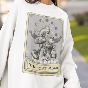 K&ouml;nnte beinhalten: Wei&szlig;es Sweatshirt mit einem grafischen Druck einer Tarotkarte. Die Karte tr&auml;gt den Titel "The Cat Mom" und zeigt eine Frau mit mehreren Katzen. Die Karte hat einen grauen Hintergrund mit goldenen Akzenten.