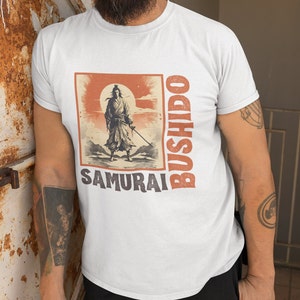 May include: White t-shirt featuring a vintage-style illustration of a samurai warrior with a sword, set against a red sun and mountain backdrop. The words "BUSHIDO" and "SAMURAI" are printed in a stylized font. The shirt is made of a soft material.