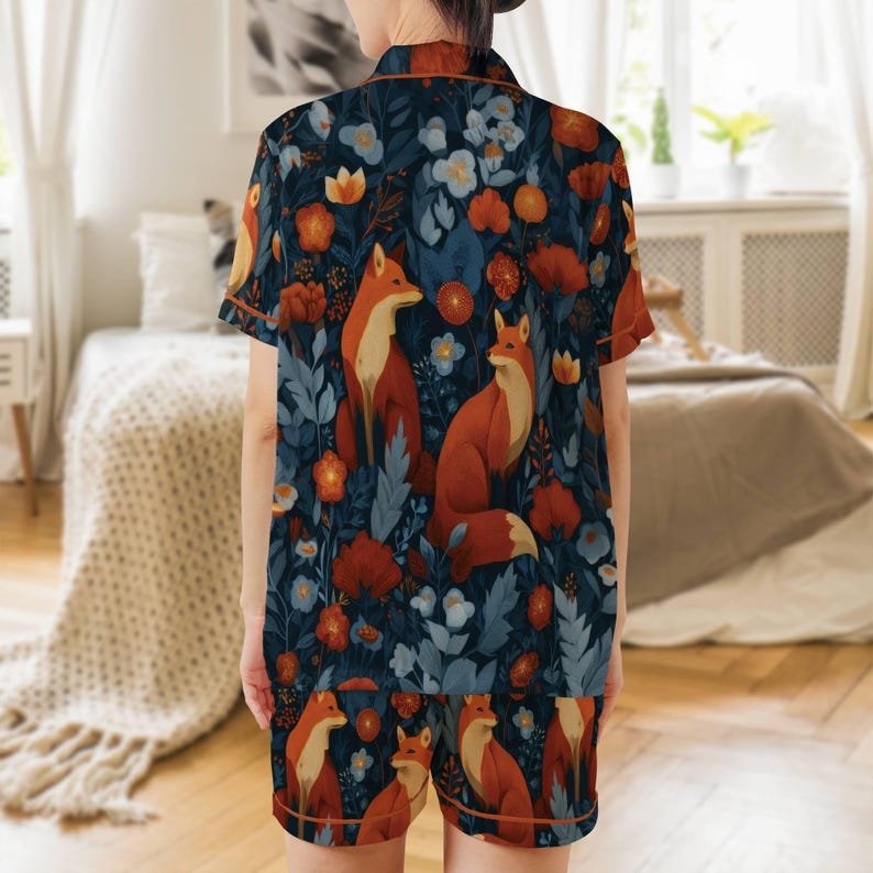 Puede incluir: Un conjunto de pijama de dos piezas con un fondo azul oscuro, adornado con un patr&oacute;n repetido de zorros, flores y follaje en tonos naranja, rojo y azul. La parte superior de manga corta y los pantalones cortos tienen ribetes naranjas.