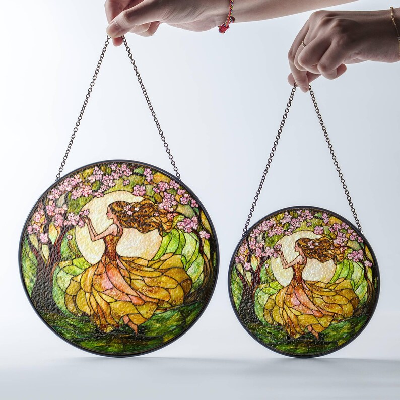 Op de afbeelding: Twee gebrandschilderde raamhangers met een vrouw in een vloeiende jurk omringd door roze bloesems en groen gebladerte. De grotere hanger heeft een diameter van 30 cm en de kleinere een diameter van 20 cm.
