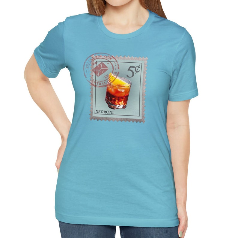 May include: A light blue t-shirt with a vintage-style postage stamp design featuring a glass of Negroni cocktail with an orange slice. The stamp reads "5&cent;" and "NEGRONI".