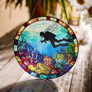 Op de afbeelding: Een rond glas-in-loodraam dat aan een ketting hangt. Het raam toont een duiker die door een kleurrijk koraalrif zwemt. De duiker is een zwart silhouet met een duikfles en luchtbellen. Het koraalrif is een levendige mix van rood, oranje, geel, groen en blauw.