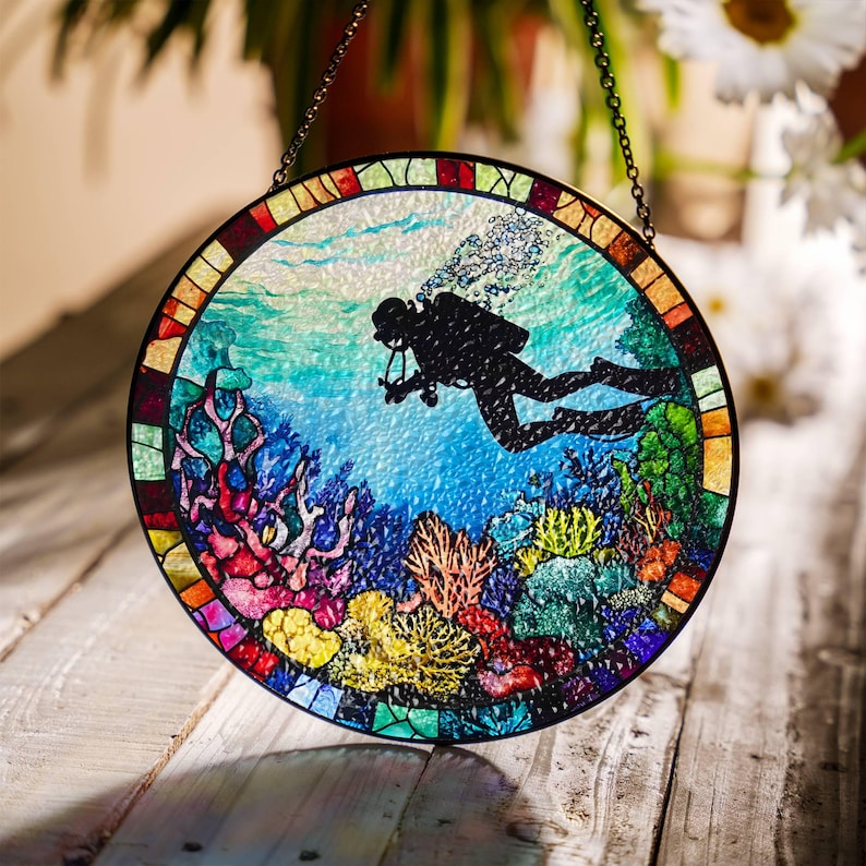 Op de afbeelding: Een rond glas-in-loodraam dat aan een ketting hangt. Het raam toont een duiker die door een kleurrijk koraalrif zwemt. De duiker is een zwart silhouet met een duikfles en luchtbellen. Het koraalrif is een levendige mix van rood, oranje, geel, groen en blauw.