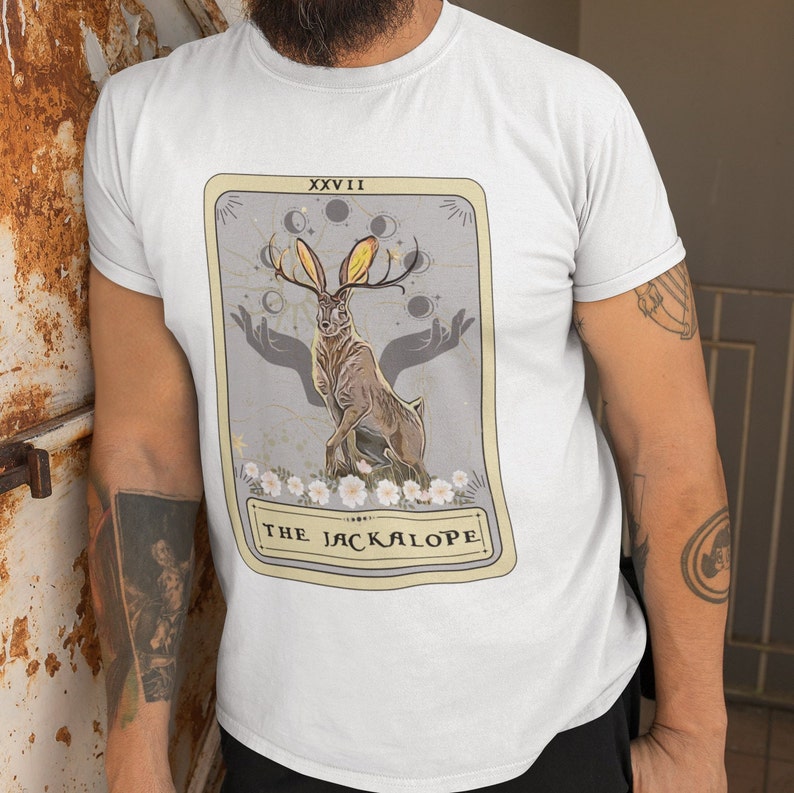 May include: A white t-shirt with a graphic of a tarot card featuring a Jackalope, a mythical creature with antlers, surrounded by a crescent moon and stars. The card is labeled "XXVII THE JACKALOPE".