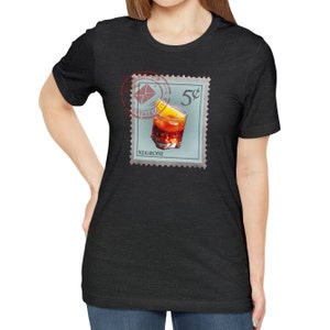 May include: A black t-shirt with a vintage-style stamp featuring a glass of Negroni with an orange slice. The stamp reads "5&cent;" and "NEGRONI".