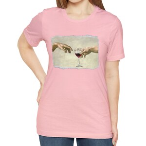 May include: A pink t-shirt with a graphic of two hands reaching towards each other, holding a glass of red wine. The graphic is in a vintage style, with a torn paper effect.