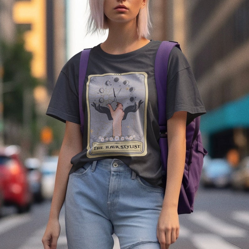 Puede incluir: Una camiseta gris oscuro con un dise&ntilde;o de carta de tarot que muestra una mano sosteniendo unas tijeras y el texto "The Hair Stylist".