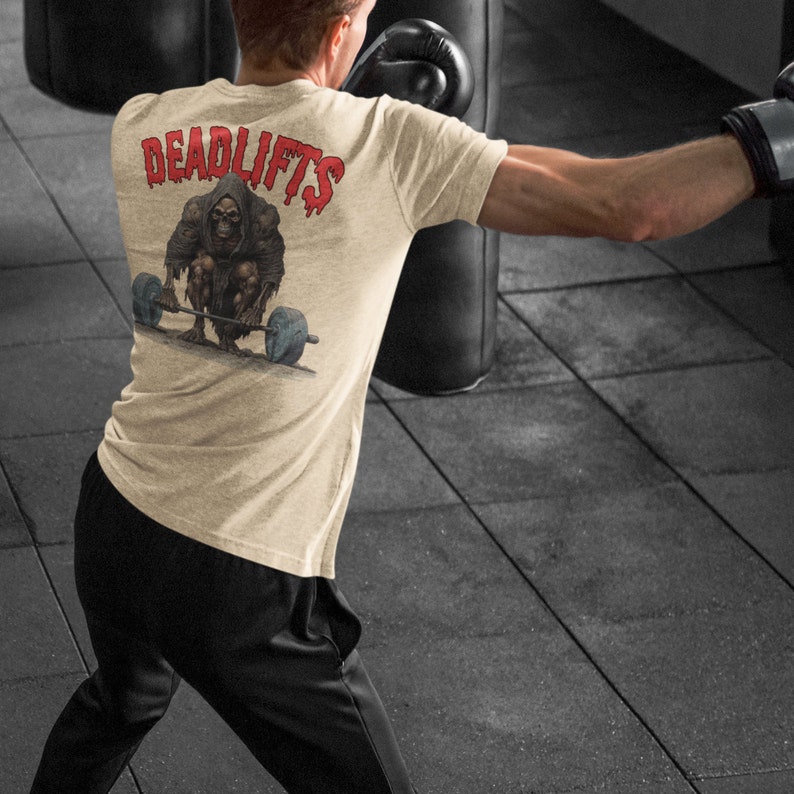 K&ouml;nnte beinhalten: Ein beiges T-Shirt mit einem Grafikdruck eines Skeletts, das eine Langhantel hebt. Der Text "DEADLIFTS" ist in Rot auf der R&uuml;ckseite des Shirts gedruckt.