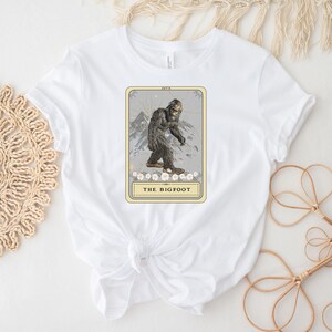 Pode incluir: Camiseta branca com um gr&aacute;fico de uma carta de tar&ocirc; com uma ilustra&ccedil;&atilde;o de Bigfoot. A carta &eacute; intitulada "The Bigfoot".