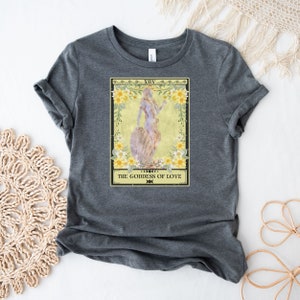 May include: A gray t-shirt with a tarot card design featuring a woman standing on a rock with flowers around her. The card reads "XIV THE GODDESS OF LOVE".