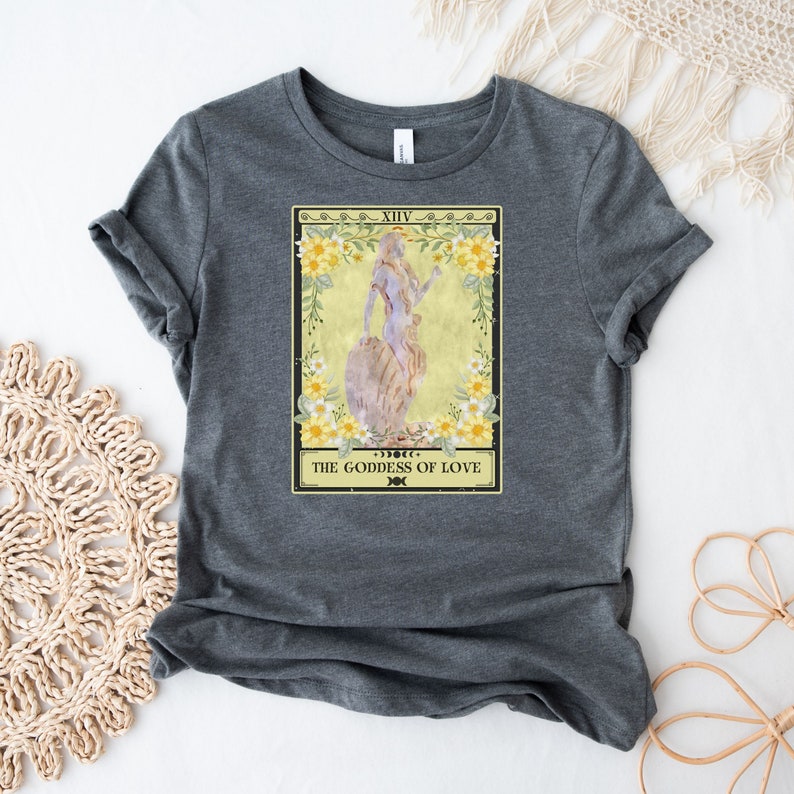 May include: A gray t-shirt with a tarot card design featuring a woman standing on a rock with flowers around her. The card reads "XIV THE GODDESS OF LOVE".