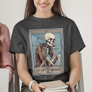 Peut inclure: Un t-shirt gris fonc&eacute; avec un motif de carte de tarot vintage repr&eacute;sentant un squelette en robe avec le texte "XVII THE OVERTHINKER".