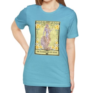 May include: A turquoise blue t-shirt with a graphic of a tarot card. The card is yellow with a floral border and features a woman standing on a rock. The card reads "XIV. THE GODDESS OF LOVE".