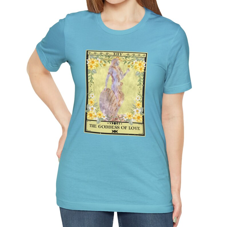 May include: A turquoise blue t-shirt with a graphic of a tarot card. The card is yellow with a floral border and features a woman standing on a rock. The card reads "XIV. THE GODDESS OF LOVE".