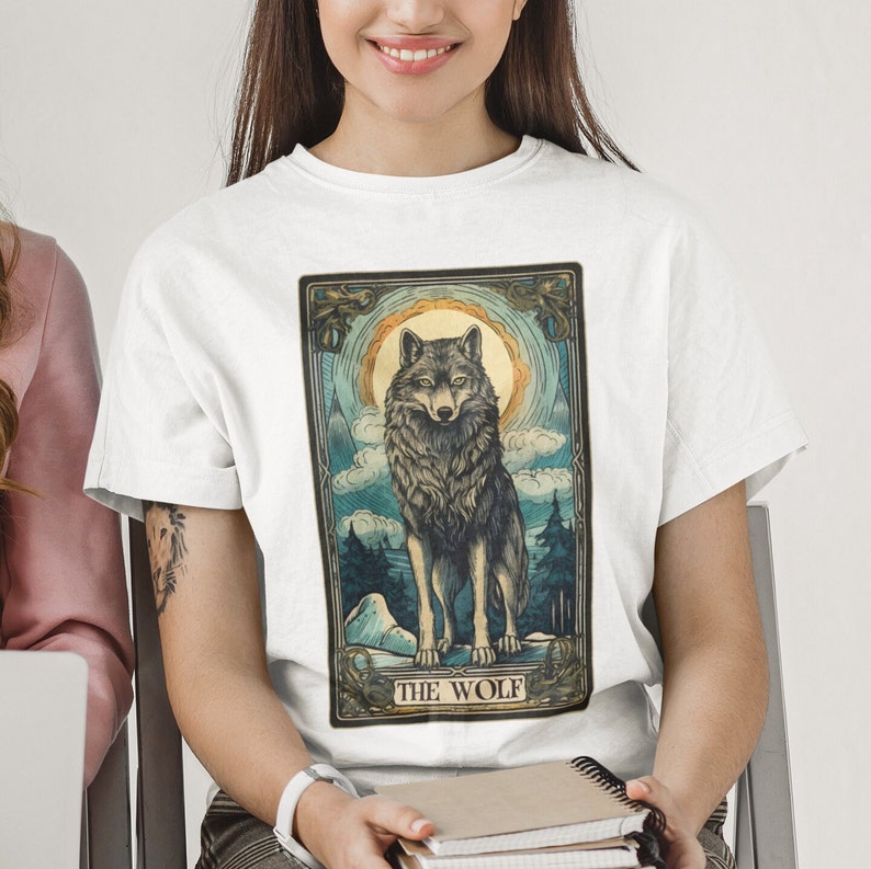 May include: White t-shirt with a vintage-style illustration of a wolf in a moonlit forest. The illustration is framed with a decorative border and includes the text "THE WOLF".