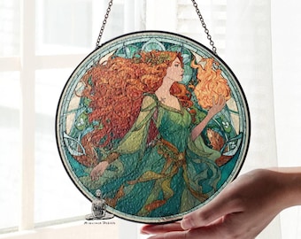 Pode incluir: Obra de arte redonda em vitral com uma mulher de cabelo ruivo e vestido verde segurando uma chama. O design inclui padrões florais e geométricos. A peça é emoldurada em um anel de metal preto e suspensa por uma corrente. Uma peça decorativa.