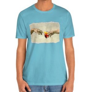 May include: A light blue t-shirt with a graphic of two hands reaching towards each other, holding a glass of red liquid with a lemon wedge. The graphic is a parody of the famous "The Creation of Adam" painting.