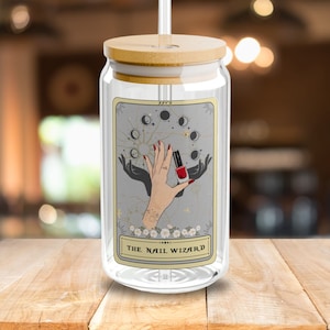 May include: A glass reusable water bottle with a bamboo lid and a straw. The bottle has a tarot card design with a hand holding a bottle of nail polish and the text "The Nail Wizard".