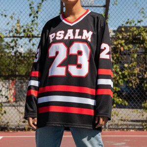 以下が含まれることがあります： 赤と白のストライプと白い文字「PSALM 23」が入った黒いジャージ。前面には数字の「23」が大きく表示されています。ジャージは赤と白のトリムが付いたVネックです。袖には白いストライプが入っています。