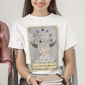May include: White t-shirt featuring a tarot card design with a woman in a nurse's uniform holding a syringe. The card is titled "La Enfermera" and has a gold border.