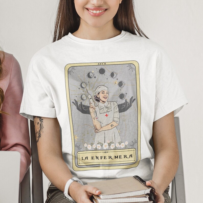 May include: White t-shirt featuring a tarot card design with a woman in a nurse's uniform holding a syringe. The card is titled "La Enfermera" and has a gold border.
