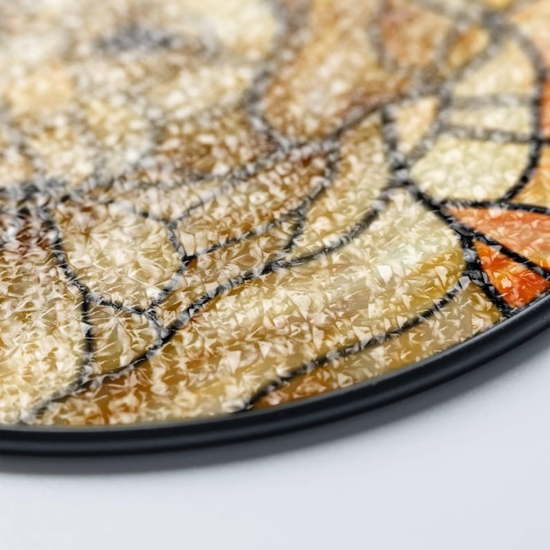 May include: Close-up of a decorative coaster with a mosaic design. The coaster features a black border and a textured surface with shades of beige, orange, and brown. Black lines outline the mosaic pattern.