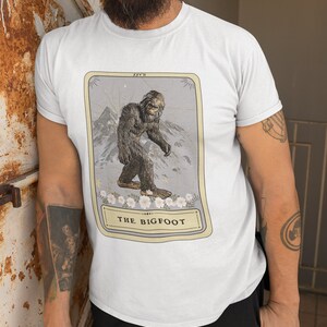 Pode incluir: Uma camiseta branca com um gr&aacute;fico de uma carta de tar&ocirc; que apresenta uma ilustra&ccedil;&atilde;o de Bigfoot. O cart&atilde;o &eacute; rotulado como "The Bigfoot" e tem o n&uacute;mero "XXV II" no canto superior direito.