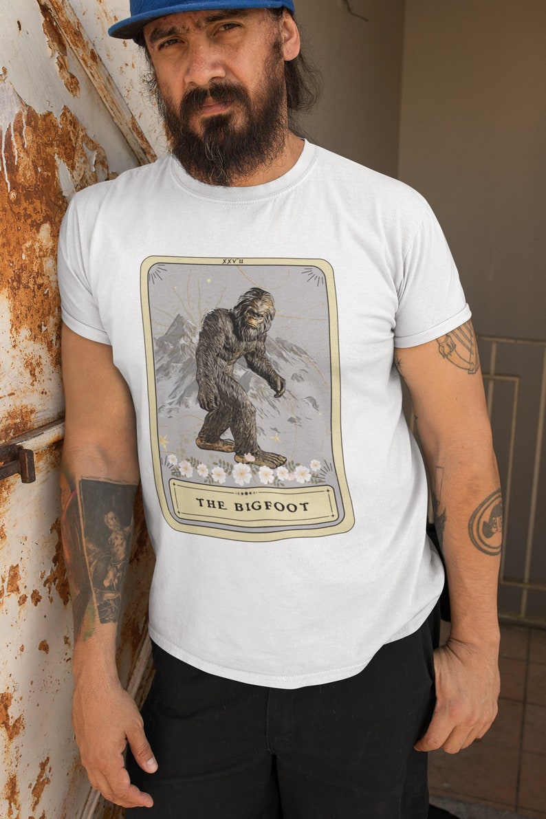 Pode incluir: Uma camiseta branca com um gr&aacute;fico de uma carta de tar&ocirc; que apresenta uma ilustra&ccedil;&atilde;o de Bigfoot. O cart&atilde;o &eacute; rotulado como "The Bigfoot" e tem o n&uacute;mero "XXV II" no canto superior direito.