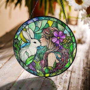 Op de afbeelding: Een hangend glas-in-loodraam toont een vrouw met lang paars haar en een wit konijn. De vrouw kust het konijn op de neus. De achtergrond is een groen en blauw glas-in-loodontwerp met bloemen.