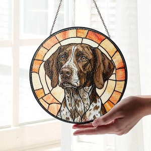 May include: Circular stained glass window art depicting a German Shorthaired Pointer dog. The dog has brown and white markings, with a detailed face. The border is a mosaic of orange and yellow hues, with a black metal frame and chain for hanging.