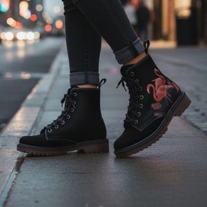 Pode incluir: Botas pretas com atacadores e sola castanha. Uma bota apresenta um desenho de flamingo rosa. As botas são usadas com jeans pretos. O fundo está desfocado, sugerindo um ambiente ao ar livre.
