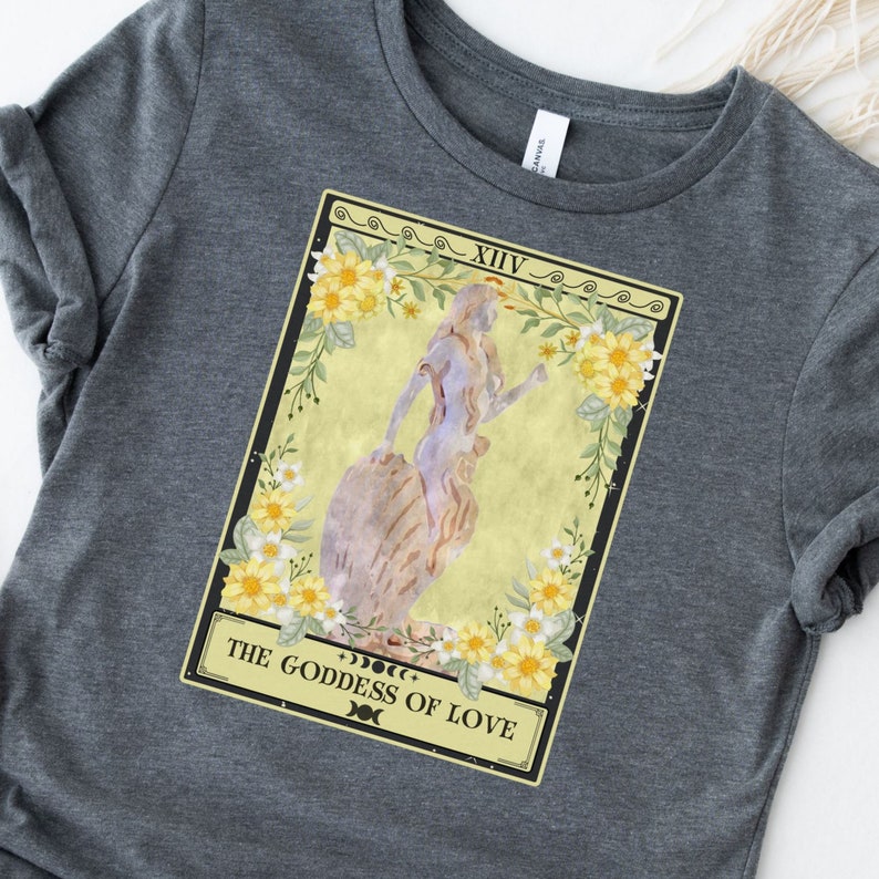 May include: A gray t-shirt with a yellow and green tarot card design. The card is labeled "XIV" and "THE GODDESS OF LOVE".