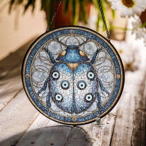 May include: Circular stained glass window art with a blue beetle design. The artwork has a dark border and a chain for hanging. The beetle has multiple eye-like details and is set against a light blue background. The diameter is approximately 20 cm.